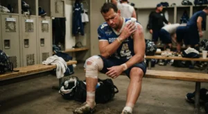 A football player wearing a blue jersey with the number 14 sits dejectedly on a wooden bench in a locker room. He is holding an ice pack to his left shoulder, which shows visible scrapes. His right leg is heavily wrapped in compression bandages from the ankle up past the knee.
