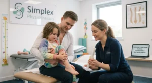 A male pediatric physiotherapist in a dark uniform crouches down to guide a young boy holding a colorful resistance rope. The session takes place in a brightly lit Simplex Physiotherapy room equipped with balance pads, colorful balls, and a children's height chart.